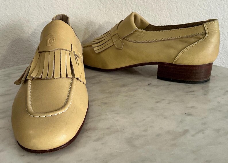 Puede incluir: Un par de mocasines de cuero amarillo claro con flecos. Los zapatos tienen una puntera redondeada, un tac&oacute;n bajo y una correa decorativa. El logotipo de Pierre Cardin es visible en el lateral de un zapato.
