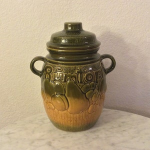 Könnte beinhalten: Eine grüne und braune Keramik-Gläser mit Deckel. Das Glas hat ein gewebtes Muster und ist mit Früchten verziert. Das Wort "Rumtopf" steht auf der Vorderseite des Glases.