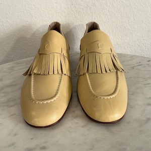 Puede incluir: Un par de mocasines de cuero amarillo claro con flecos. Los zapatos tienen una punta redondeada y un tac&oacute;n peque&ntilde;o. El logotipo de Pierre Cardin est&aacute; grabado en la parte superior de cada zapato. Las suelas son de color marr&oacute;n oscuro.
