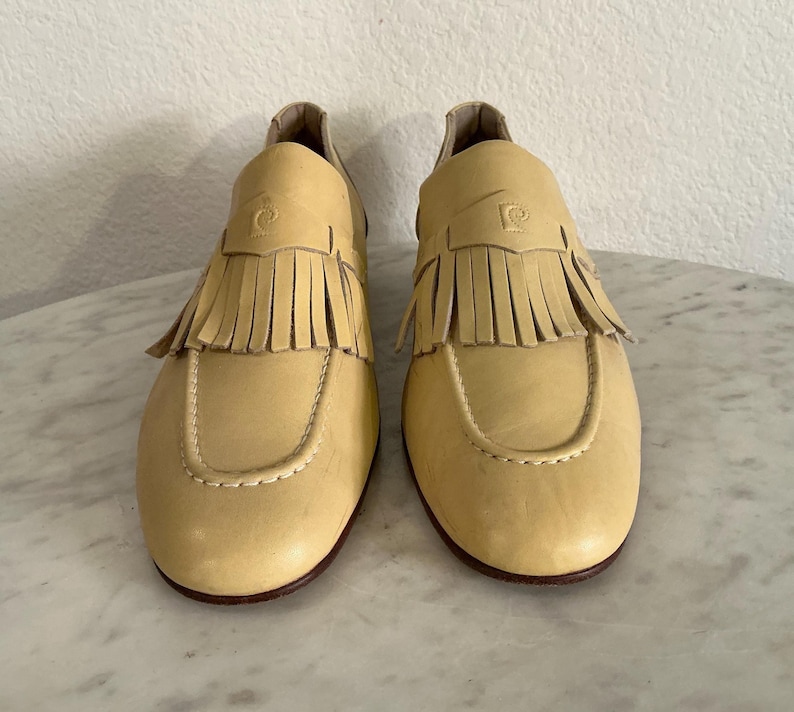 Puede incluir: Un par de mocasines de cuero amarillo claro con flecos. Los zapatos tienen una punta redondeada y un tac&oacute;n peque&ntilde;o. El logotipo de Pierre Cardin est&aacute; grabado en la parte superior de cada zapato. Las suelas son de color marr&oacute;n oscuro.