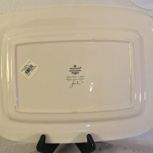 Puede incluir: Plato rectangular blanco para servir con un borde elevado y bordes festoneados. El plato tiene una pegatina en la parte inferior con el texto "WEDGWOOD&reg; SARAH'S GARDEN" y una firma manuscrita.