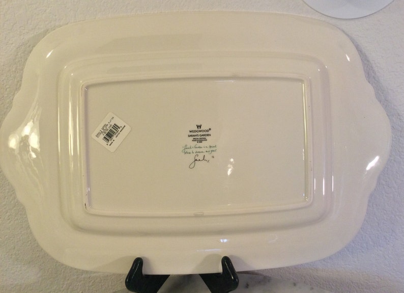 Puede incluir: Plato rectangular blanco para servir con un borde elevado y bordes festoneados. El plato tiene una pegatina en la parte inferior con el texto "WEDGWOOD&reg; SARAH'S GARDEN" y una firma manuscrita.