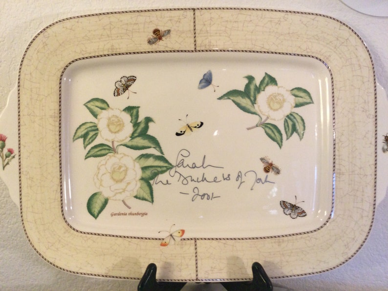 Puede incluir: Un plato de servir de cer&aacute;mica blanca con un dise&ntilde;o floral con camelias blancas y hojas verdes. El plato tiene un borde marr&oacute;n con un dise&ntilde;o de cuerda. El texto "Sarah The Duchess of York - 2001" est&aacute; escrito con tinta negra en el plato.