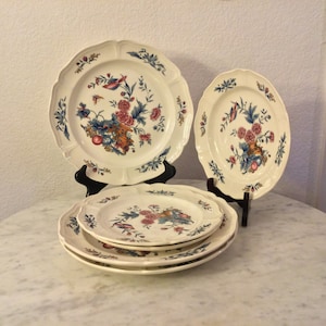 Puede incluir: Un conjunto de platos de cerámica color crema con un diseño floral y de pájaros. Los platos tienen un borde festoneado y están decorados con flores y pájaros rosas, azules y naranjas. Los platos se exhiben sobre soportes negros.