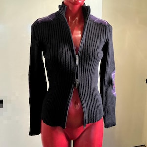 Op de afbeelding: Een zwarte geribbelde gebreide trui met ritssluiting en paarse ruitaccenten op de schouders en ellebogen. De trui heeft een hoge kraag en een zilveren rits. Gepresenteerd op een mannequin.