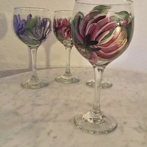 Peut inclure: Quatre verres &agrave; vin en verre transparent avec des motifs floraux peints. Les verres ont un bol rond et une longue tige. Les fleurs sont peintes dans des tons de rose, violet et vert.
