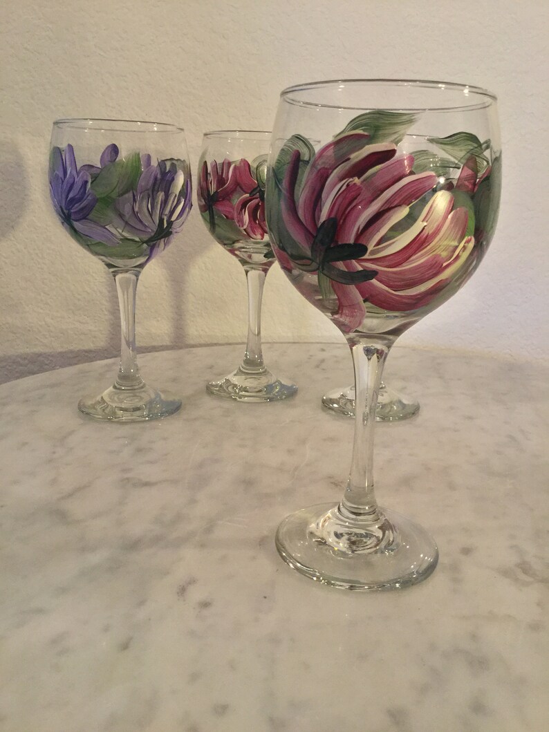 Peut inclure: Quatre verres &agrave; vin en verre transparent avec des motifs floraux peints. Les verres ont un bol rond et une longue tige. Les fleurs sont peintes dans des tons de rose, violet et vert.