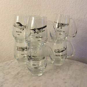 Könnte beinhalten: Sechs durchsichtige Glasbecher mit Schwarzweißbildern von Oldtimer-Flugzeugen und ihren Namen und Jahren. Die Becher stehen auf einer weißen Marmorfläche.