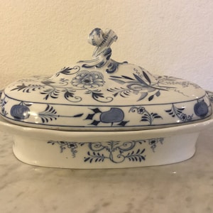 Puede incluir: Un plato de servir de porcelana blanca con un diseño floral azul y blanco. El plato tiene una tapa con un pomo decorativo.