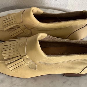 Puede incluir: Un par de mocasines de cuero color beige claro. Estos zapatos tienen un dise&ntilde;o cl&aacute;sico, con punta redondeada, flecos decorativos y un peque&ntilde;o detalle de correa. Est&aacute;n sobre un tac&oacute;n bajo. Los zapatos parecen sin usar.