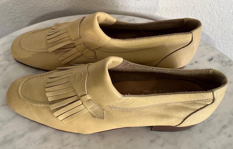 Puede incluir: Un par de mocasines de cuero color beige claro. Estos zapatos tienen un dise&ntilde;o cl&aacute;sico, con punta redondeada, flecos decorativos y un peque&ntilde;o detalle de correa. Est&aacute;n sobre un tac&oacute;n bajo. Los zapatos parecen sin usar.