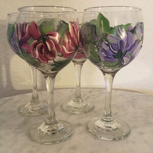 Peut inclure: Quatre verres &agrave; vin en verre transparent avec des motifs floraux peints &agrave; la main. Deux verres pr&eacute;sentent des fleurs roses avec des feuilles vertes, et deux verres pr&eacute;sentent des fleurs violettes avec des feuilles vertes.