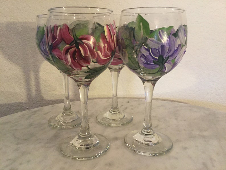 Peut inclure: Quatre verres &agrave; vin en verre transparent avec des motifs floraux peints &agrave; la main. Deux verres pr&eacute;sentent des fleurs roses avec des feuilles vertes, et deux verres pr&eacute;sentent des fleurs violettes avec des feuilles vertes.