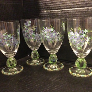 May include: Four clear glass wine glasses with green stems and bases. Each glass is hand-painted with a cluster of blue and green flowers.