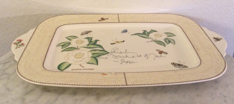 Puede incluir: Una bandeja rectangular para servir con fondo beige y borde decorativo. La bandeja presenta un dise&ntilde;o floral con camelias blancas, hojas verdes y mariposas. El texto "Each Journey is Part of the Story" est&aacute; escrito en cursiva en la bandeja.