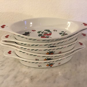 Może przedstawiać: Zestaw czterech owalnych ceramicznych naczyń do pieczenia w kolorze białym z zielonymi i różowymi wzorami kwiatowymi. Naczynia mają uchwyty po obu stronach i przedstawiają różne wzory owoców, w tym wiśnie, gruszki i pomarańcze.