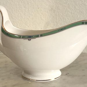 Könnte beinhalten: Weiße Porzellan-Sauciere mit gebogenem Griff und Ausguss. Der Rand ist mit einem grünen und blauen geometrischen Muster verziert, und goldfarbene Zierleisten akzentuieren den Griff und den Sockel. Ein klassisches Servierstück.