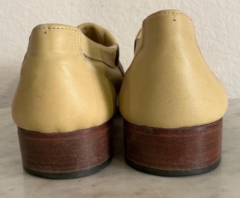 Puede incluir: Un par de zapatos de cuero de color beige claro con tacones marrones oscuros. Los zapatos tienen una punta redondeada y un tac&oacute;n bajo. El cuero parece liso y los zapatos est&aacute;n en buen estado.