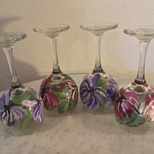 Peut inclure: Quatre verres &agrave; vin peints &agrave; la main avec des motifs floraux. Chaque verre pr&eacute;sente un arrangement diff&eacute;rent de fleurs violettes et roses avec des feuilles vertes.
