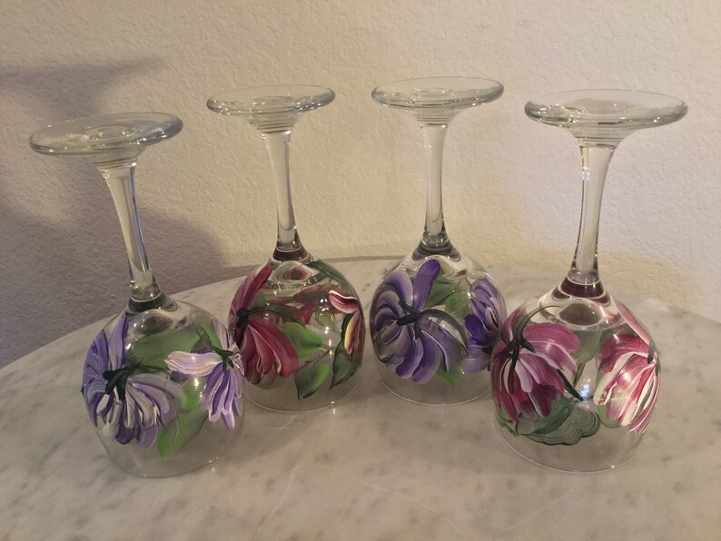 Peut inclure: Quatre verres &agrave; vin peints &agrave; la main avec des motifs floraux. Chaque verre pr&eacute;sente un arrangement diff&eacute;rent de fleurs violettes et roses avec des feuilles vertes.