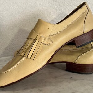 Puede incluir: Un par de mocasines de cuero amarillo claro con detalles de borlas. Los zapatos tienen un tac&oacute;n bajo y una suela marr&oacute;n. Las costuras son visibles a lo largo de los bordes del zapato.