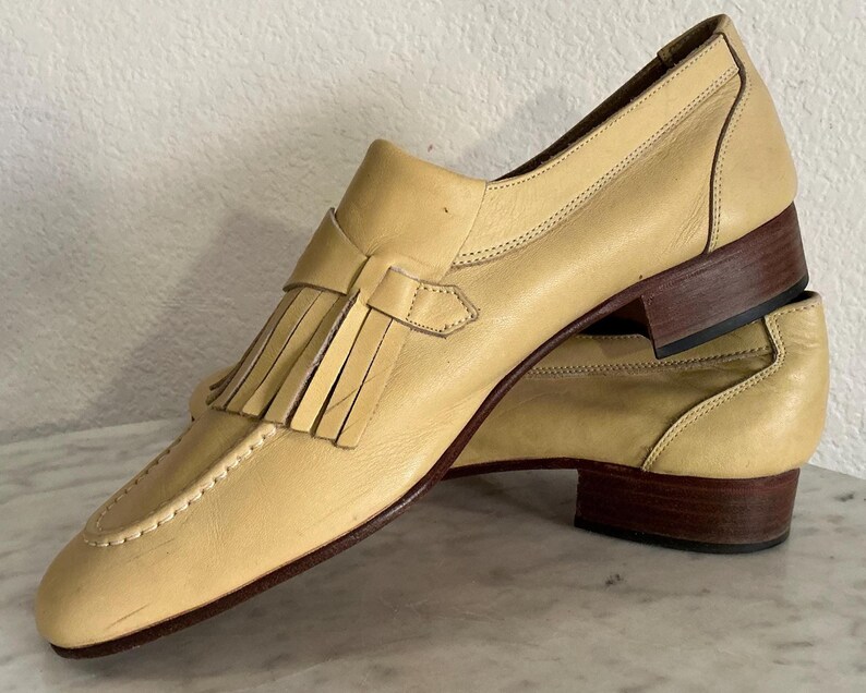 Puede incluir: Un par de mocasines de cuero amarillo claro con detalles de borlas. Los zapatos tienen un tac&oacute;n bajo y una suela marr&oacute;n. Las costuras son visibles a lo largo de los bordes del zapato.
