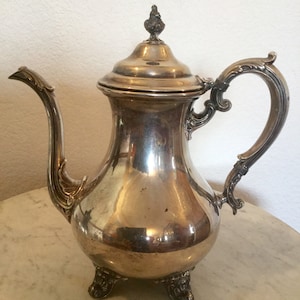 May include: A silver teapot with a round body, a curved spout, and a decorative handle. The teapot has three feet and a lid with a finial.
