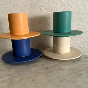 May include: Two modern candle holders. One has an orange cylinder on a yellow disc, atop a blue cylinder and disc. The other has a teal cylinder on a green disc, atop a cream cylinder and disc. Decorative home decor.