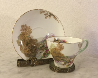 May include: A vintage teacup and saucer set with a delicate floral and landscape design. The cup and saucer are white with gold trim and feature a painted scene of a bridge over a stream in a wooded area.