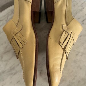 Puede incluir: Un par de zapatos de cuero de color beige claro con tac&oacute;n bajo. Cada zapato presenta un detalle de flecos decorativos y un dise&ntilde;o cosido a lo largo de los lados. Los tacones son de color marr&oacute;n oscuro.