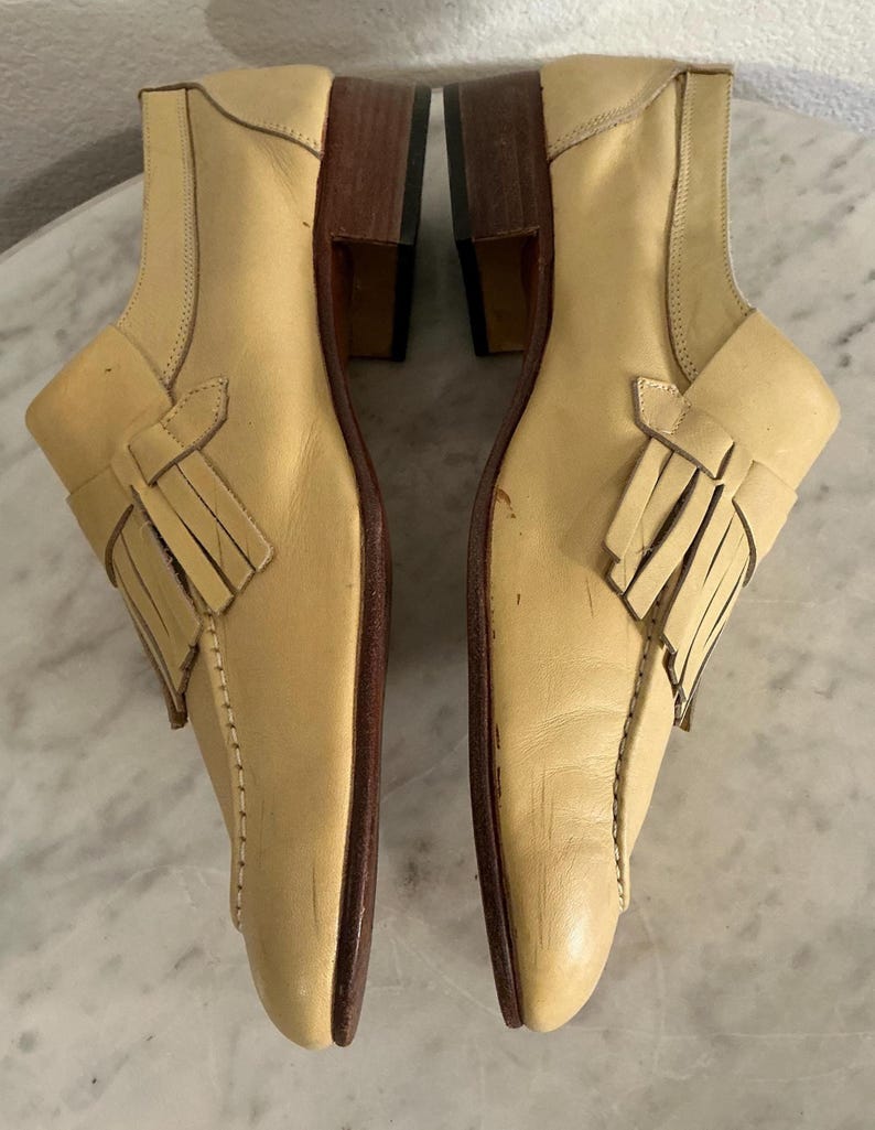 Puede incluir: Un par de zapatos de cuero de color beige claro con tac&oacute;n bajo. Cada zapato presenta un detalle de flecos decorativos y un dise&ntilde;o cosido a lo largo de los lados. Los tacones son de color marr&oacute;n oscuro.