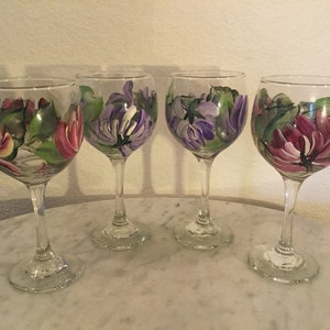 Peut inclure: Ensemble de quatre verres &agrave; vin peints &agrave; la main avec des motifs floraux. Deux verres pr&eacute;sentent des fleurs roses avec des feuilles vertes, tandis que les deux autres verres pr&eacute;sentent des fleurs violettes avec des feuilles vertes.