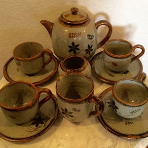May include: A set of six ceramic teacups and saucers with a brown and grey speckled glaze. The cups and saucers feature a floral design in brown, orange, and green. The set also includes a teapot with a matching design.