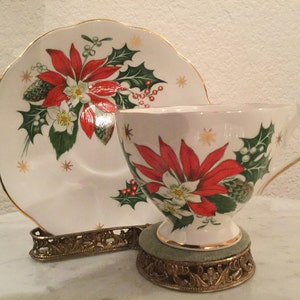 May include: A white teacup and saucer with a Christmas poinsettia design. The cup and saucer have a gold trim and are decorated with red poinsettias, green holly leaves, and white flowers. The saucer has a scalloped edge.