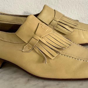 Puede incluir: Un par de mocasines de cuero amarillo claro con flecos y un tac&oacute;n peque&ntilde;o. Los zapatos tienen un dise&ntilde;o cl&aacute;sico con costuras decorativas alrededor de la puntera y una correa de cuero en la parte superior.