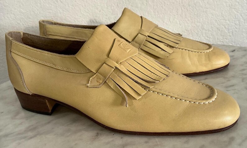 Puede incluir: Un par de mocasines de cuero amarillo claro con flecos y un tac&oacute;n peque&ntilde;o. Los zapatos tienen un dise&ntilde;o cl&aacute;sico con costuras decorativas alrededor de la puntera y una correa de cuero en la parte superior.