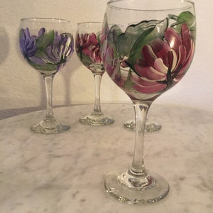 Peut inclure: Quatre verres &agrave; vin peints &agrave; la main avec des motifs floraux. Les verres sont transparents et ont un bol rond et une longue tige. Les motifs sont dans des tons de violet, rose et vert.