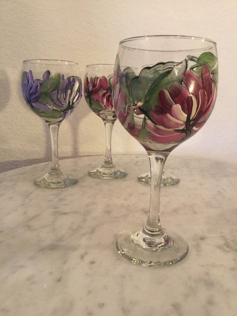 Peut inclure: Quatre verres &agrave; vin peints &agrave; la main avec des motifs floraux. Les verres sont transparents et ont un bol rond et une longue tige. Les motifs sont dans des tons de violet, rose et vert.