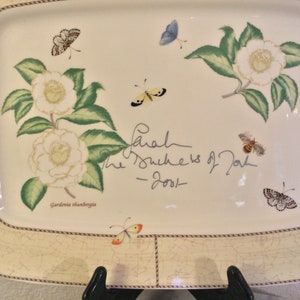 Puede incluir: Una bandeja de servir de cer&aacute;mica blanca con un dise&ntilde;o floral que presenta flores de camelia blancas, hojas verdes y mariposas. La bandeja tiene un borde de cuerda marr&oacute;n y una firma que dice "Sarah The Duchess of York - 2001 -" y el texto "Gardenia thunbergia".