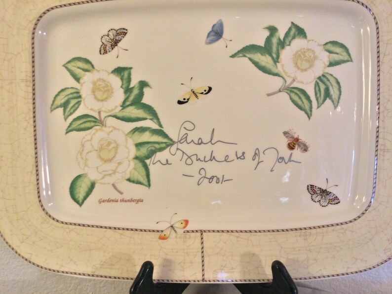 Puede incluir: Una bandeja de servir de cer&aacute;mica blanca con un dise&ntilde;o floral que presenta flores de camelia blancas, hojas verdes y mariposas. La bandeja tiene un borde de cuerda marr&oacute;n y una firma que dice "Sarah The Duchess of York - 2001 -" y el texto "Gardenia thunbergia".