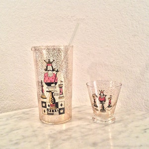 Può includere: Un set di due shaker per cocktail in vetro trasparente con una superficie testurizzata. Lo shaker più grande presenta un design di un pezzo di scacchi del re con altri pezzi di scacchi intorno. Lo shaker più piccolo ha lo stesso design ma è più piccolo. Entrambi gli shaker hanno uno schema di colori oro e rosa.