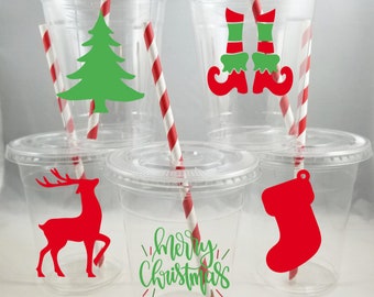 May include: Five clear plastic cups with red and white striped straws. Each cup has a different Christmas-themed design: a green Christmas tree, red elf shoes, a red reindeer, a green "Merry Christmas" message, and a red stocking.