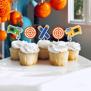 May include: Six cupcakes decorated with colorful toppers featuring a target, crossed darts, and toy guns. The cupcakes are frosted with white frosting and are arranged on a white cake stand.