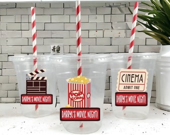 May include: Three clear plastic cups with red and white striped straws. Each cup has a different movie-themed design: a clapperboard, a box of popcorn, and a movie ticket. The cups are labeled "Darryl's Movie Night!"