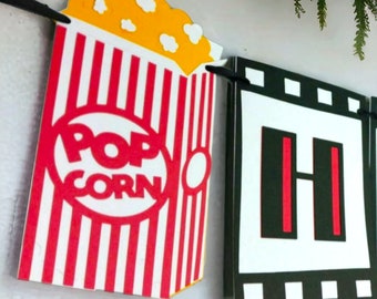May include: Red and white striped popcorn box with the words "POP CORN" in a red circle.  The box is part of a movie themed banner with a black and white film strip.