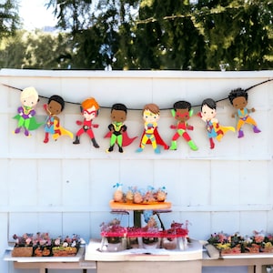 May include: A colorful superhero party banner with eight characters wearing capes and masks. The banner is strung on a white wall with a dessert table in the foreground.