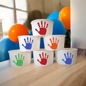 May include: Six white paper cups stacked in a pyramid shape. Each cup has a different colored handprint on the front: red, orange, blue, green, and purple. The cups are on a wooden surface.