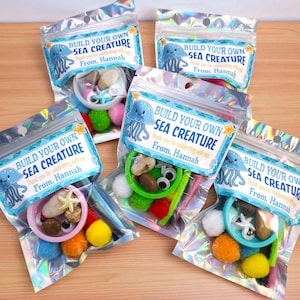 May include: Five clear, iridescent bags filled with craft supplies for creating sea creatures. Each bag includes a label with the text "Build Your Own Sea Creature" and a jellyfish illustration. Contents include pom-poms, googly eyes, and small stones.
