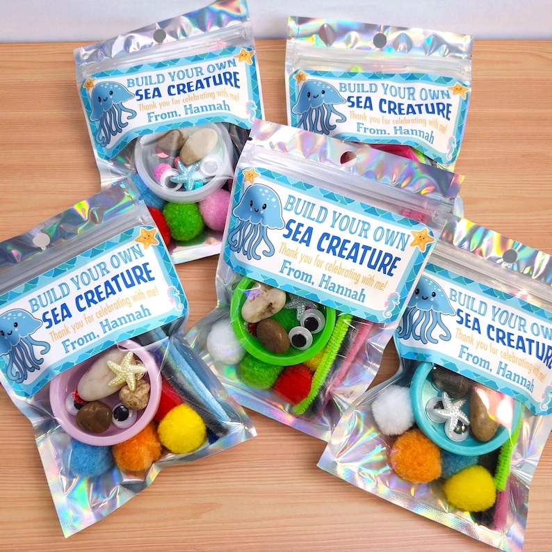May include: Five clear, iridescent bags filled with craft supplies for creating sea creatures. Each bag includes a label with the text "Build Your Own Sea Creature" and a jellyfish illustration. Contents include pom-poms, googly eyes, and small stones.
