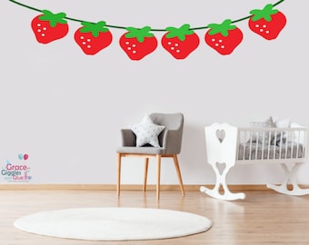 May include: A white crib with a heart-shaped cutout on the headboard, a gray chair with a star-shaped pillow, and a white rug in a nursery setting. A red and green strawberry garland is on the wall with the text "Grace-Giggles and Glue" below it.